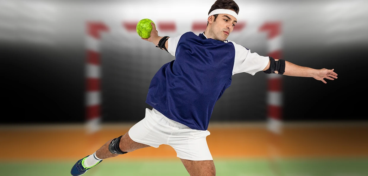 Un joueur de handball en mouvement, vêtu d'un maillot bleu, d'un short blanc et de protège-bras, s'apprête à lancer une balle verte vers un filet de but flou dans une salle omnisports.